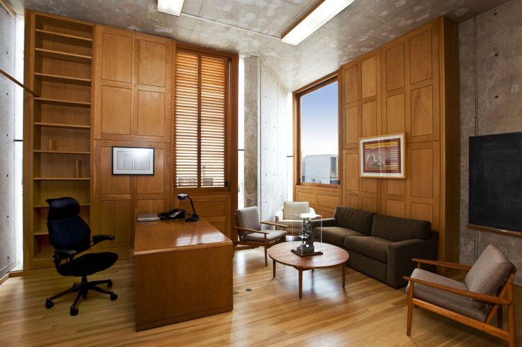 Oficina moderna con paredes de cemento y madera, equipado con un escritorio, sillas, y amplias ventanas que permiten la entrada de luz natural.