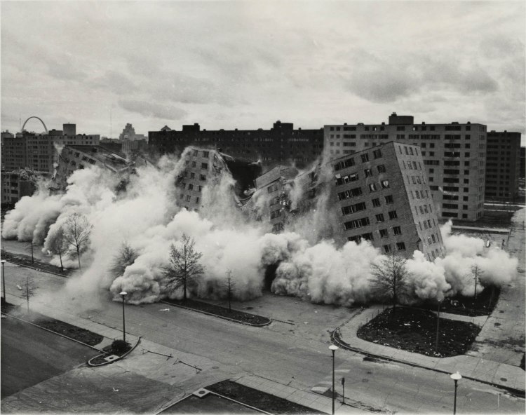 A historical black and white photograph capturing the demolition of the Pruitt-Igoe housing project, with large clouds of dust and debris rising as the buildings collapse.