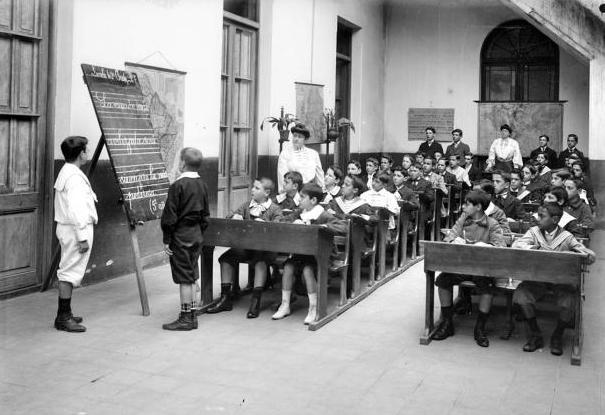 Historical black and white photograph of a classroom, featuring a teacher and a group of students seated at wooden desks, with one student standing at a chalkboard.