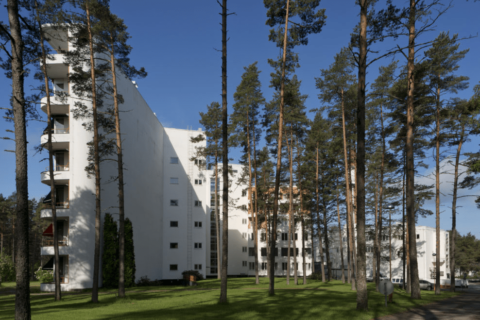 A modern white building surrounded by tall pine trees, with balconies visible on the left side under a clear blue sky.