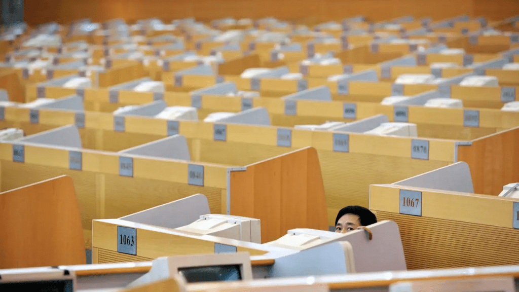 A young professional peeks over the divider of a row of identical cubicles in a vast, empty office space.