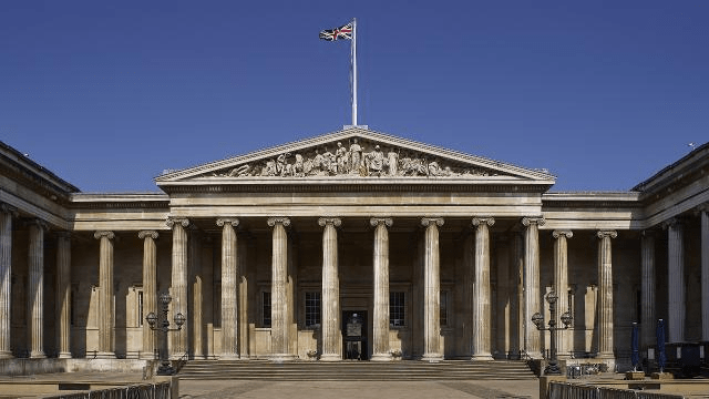 Front view of a historic building featuring a classic portico with multiple columns, a triangular pediment, and a flag atop the structure under a clear blue sky.