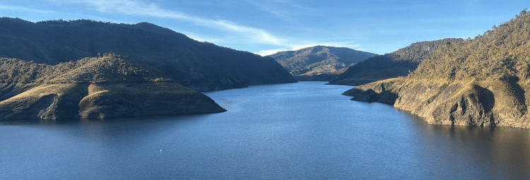 A panoramic view of a serene reservoir surrounded by rolling mountains and a clear blue sky.