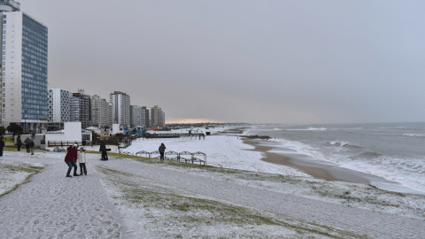 A winter beach scene showing a snowy shoreline and people walking along a sandy path next to a coast with waves crashing, buildings in the background under a gray sky.