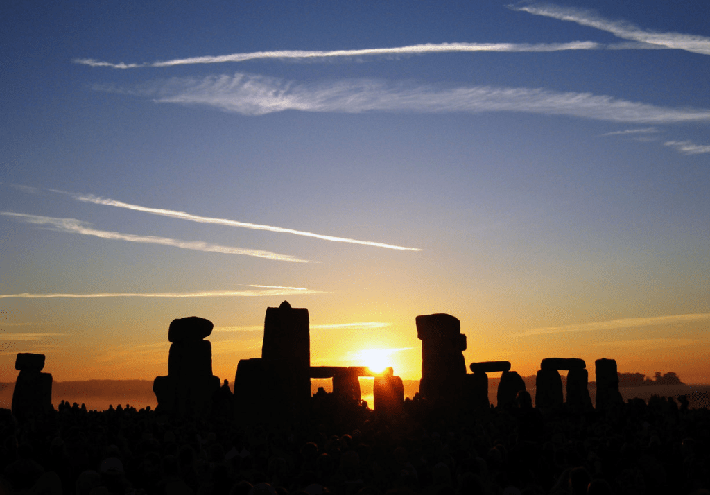 Amanecer en Stonehenge, con personas reunidas frente a las grandes piedras mientras el sol sale en el horizonte, iluminando el cielo en tonos cálidos.