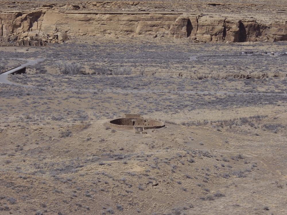 Vista aérea de un complejo ancestral en Cañón del Chaco, mostrando un gran kiva circular en un paisaje árido, con un camino serpenteante en el fondo.