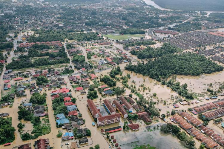 Vista aérea de una ciudad inundada, mostrando calles y edificios parcialmente sumergidos en agua, con áreas verdes y una carretera en el fondo.