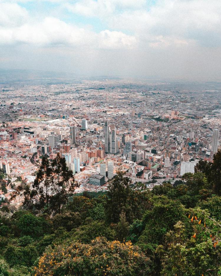 Panoramic view of Bogotá, Colombia, showcasing the city's urban landscape surrounded by lush greenery and distant mountains under a partly cloudy sky.