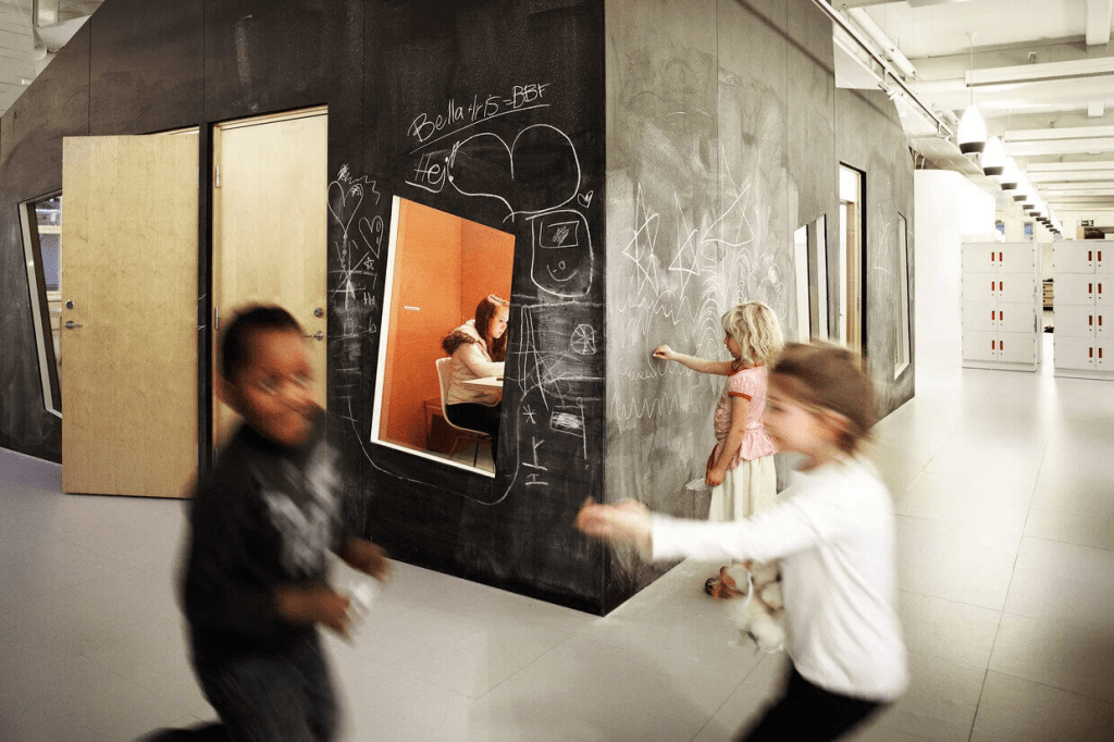 Niños jugando en un entorno escolar moderno con paredes de pizarra y puertas de madera, donde uno de ellos está escribiendo en la pizarra mientras otro corre.