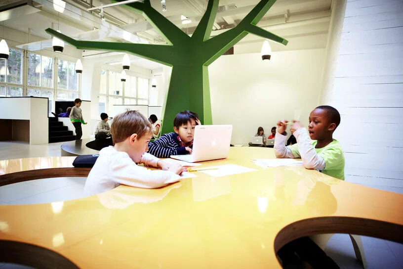 Tres niños sentados en una mesa amarilla en un aula moderna, con un gran árbol verde de fondo. Un niño trabaja en una computadora portátil mientras los otros escriben en hojas de papel.