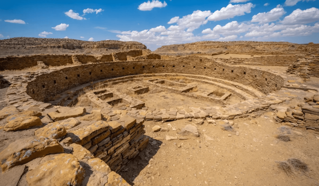 Vista del Gran Kiva en el Cañón del Chaco, un lugar de reunión ceremonial construido con piedras naturales en una estructura circular, rodeado de desierto y montañas bajo un cielo azul con nubes.