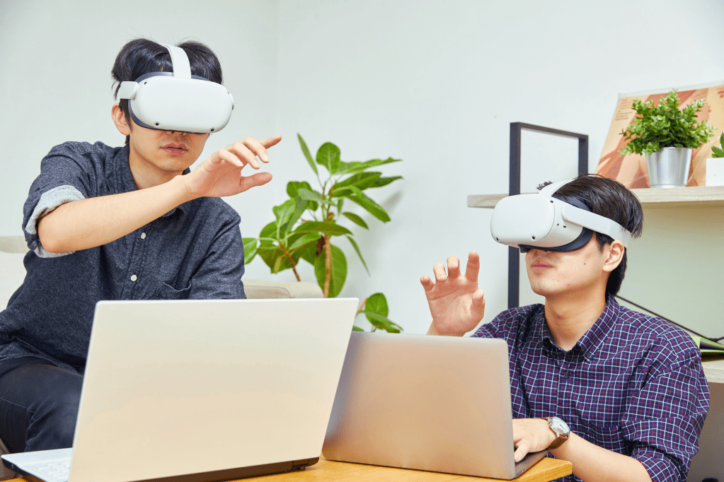 Two individuals wearing virtual reality headsets are seated at a table, engaging with laptops while reaching out with their hands, indicating interaction with a digital environment.