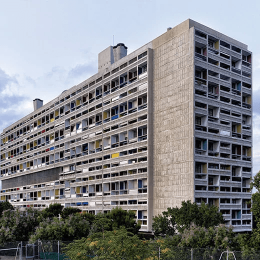 Exterior view of a Brutalist residential building with concrete façade and colorful window frames, surrounded by greenery.