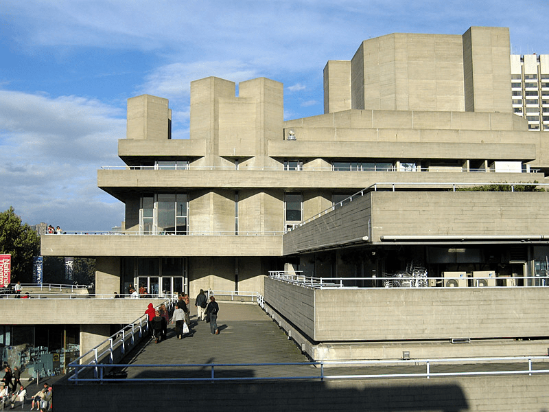 Exterior view of a Brutalist architecture building with raw concrete surfaces and angular forms, featuring a pathway where people are walking.
