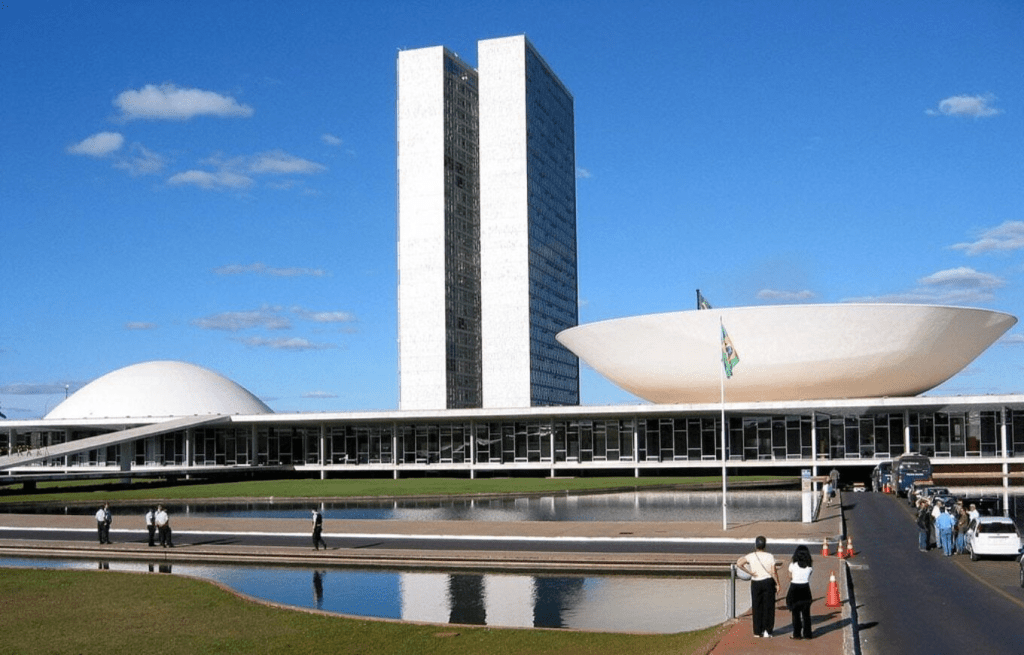 A view of the National Congress of Brazil, showcasing its modernist architecture with distinctive curved and rectangular shapes, surrounded by a blue sky and green landscape, with people walking near a reflecting pool.