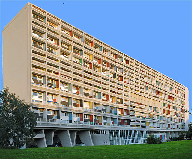 A large concrete residential building with multiple balconies and colorful window panels, set against a clear blue sky.