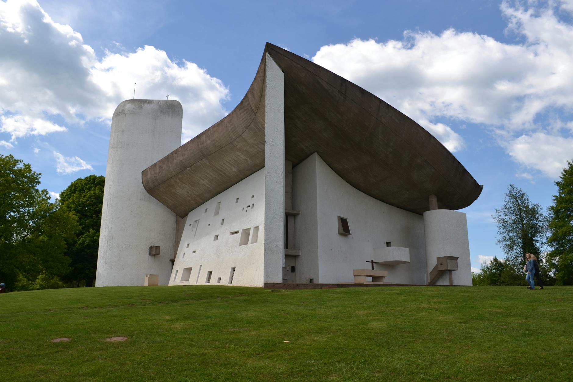 A modern Brutalist building featuring a concrete cantilevered roof, angular shapes, and a white facade set against a blue sky with fluffy clouds.