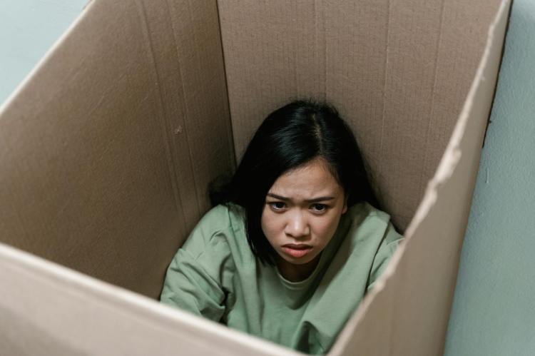 A young woman in a green sweater sitting inside a cardboard box, looking anxious and distressed.