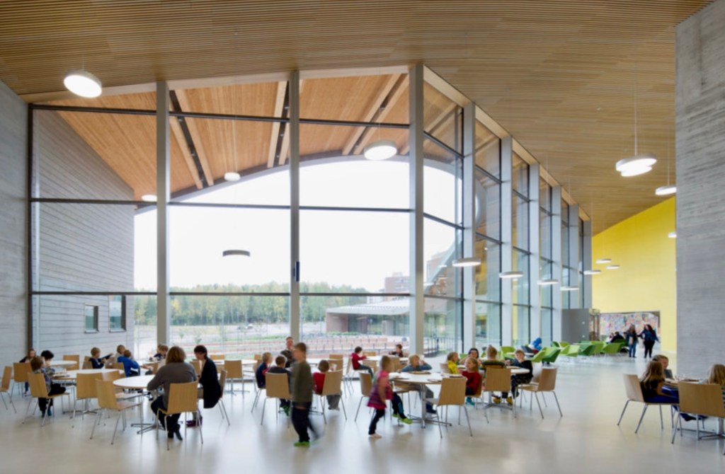 Interior view of a modern classroom with large windows, wooden beams, and students actively engaged in learning at tables.