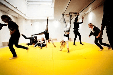 Children engaged in physical activities like handstands and play on a bright yellow mat in a spacious indoor setting.