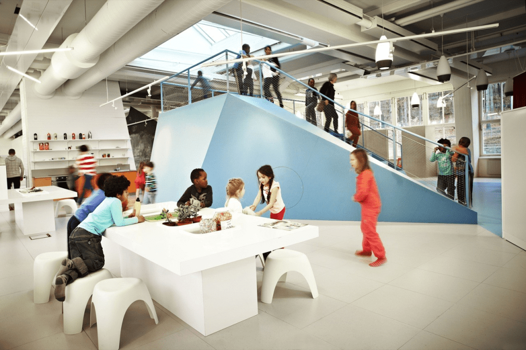 A bright and open classroom featuring children engaged in various activities, with some sitting at white tables and others ascending a blue ramp. The space is designed for flexibility and movement, emphasizing an interactive learning environment.
