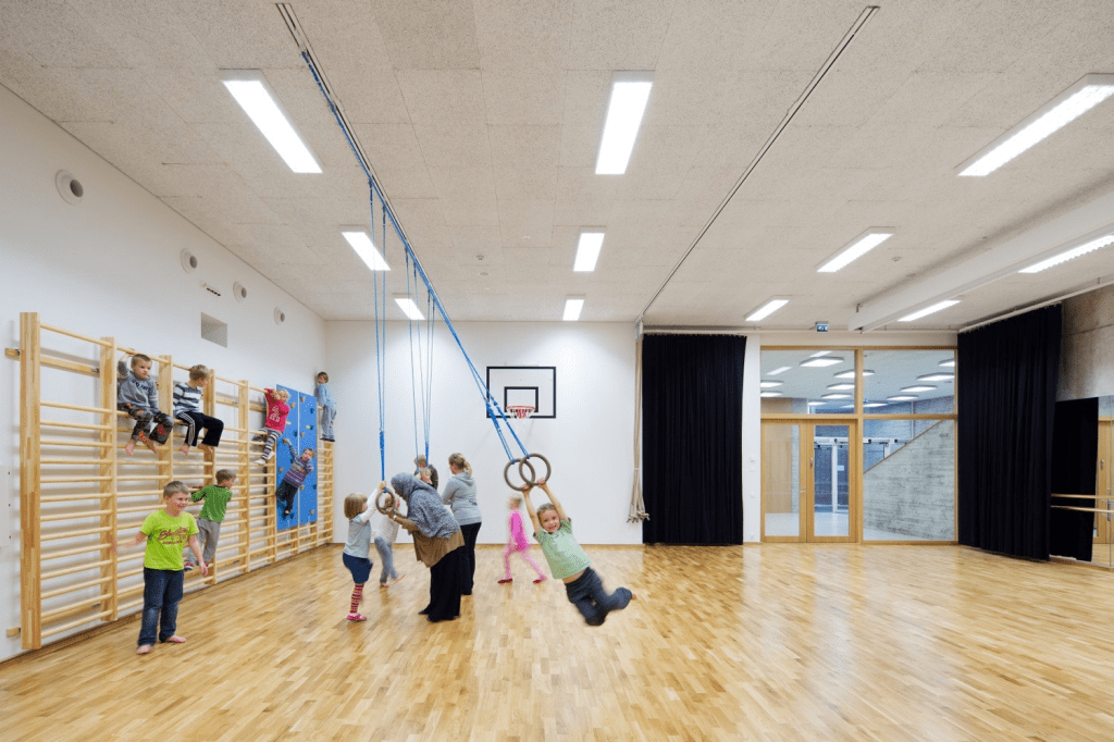 A group of children engaged in physical activities in a brightly lit classroom equipped with climbing walls and gymnastic rings.