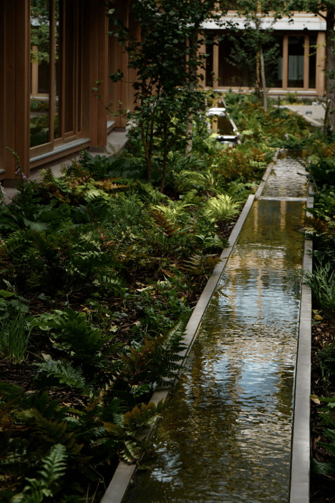 Jardín interior del Appleby Blue Almshouse, con un canal de agua reflejando el entorno y rodeado de plantas verdes y árboles.
