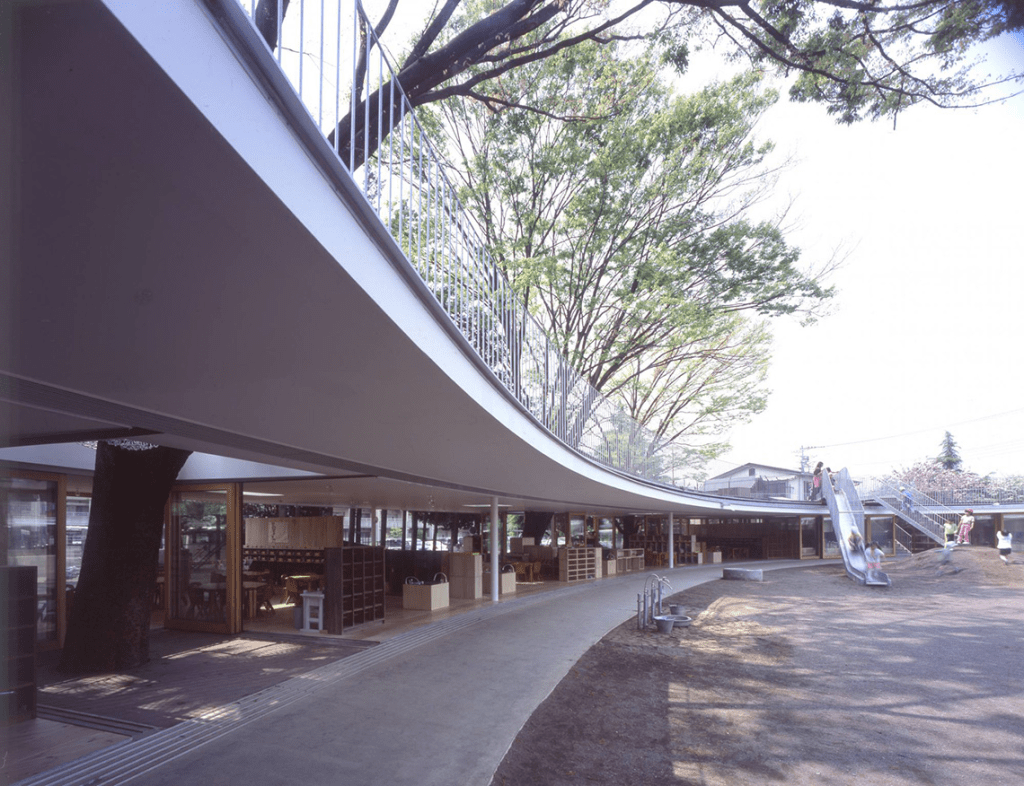 A modern school building with a curved roof design, integrated with trees, featuring a playground with a slide and open outdoor spaces for children to explore.