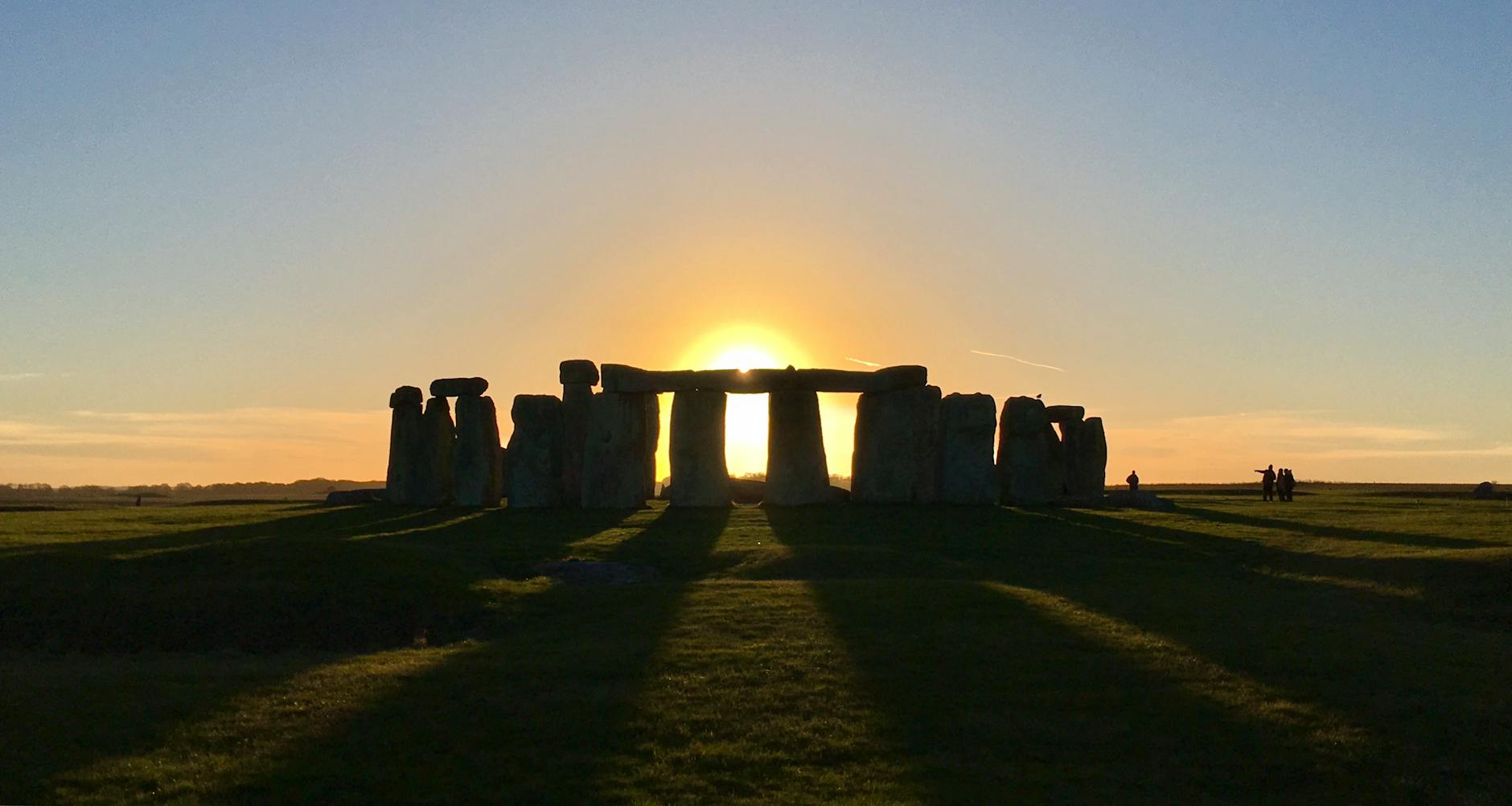 A silhouette of Stonehenge at sunset, with sunlight shining behind the ancient stones and casting long shadows on the grass.