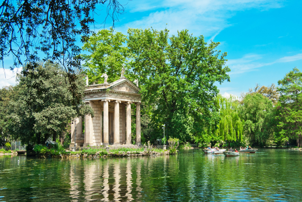 A picturesque scene featuring a classical-style building with columns beside a serene lake, surrounded by lush green trees and blue skies. Small boats can be seen floating on the water.