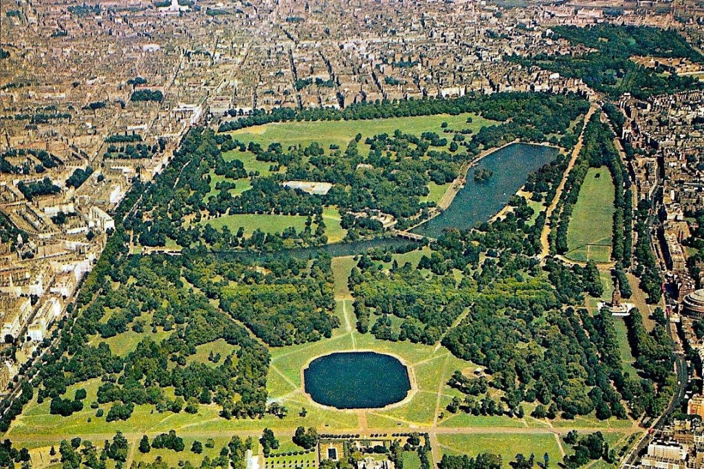 Aerial view of a large urban park surrounded by city buildings, featuring lush greenery, a pond, and open grassy areas.