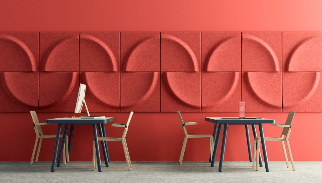 Una sala de trabajo moderna con una pared roja decorada con paneles acústicos texturizados en tonos similares, y dos mesas con sillas, un ordenador en una de ellas.