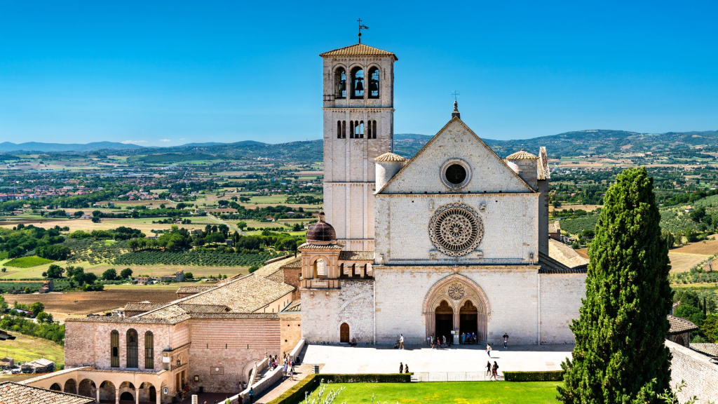 View of the Basilica of Saint Francis in Assisi, Italy, surrounded by lush greenery and scenic hills under a clear blue sky.