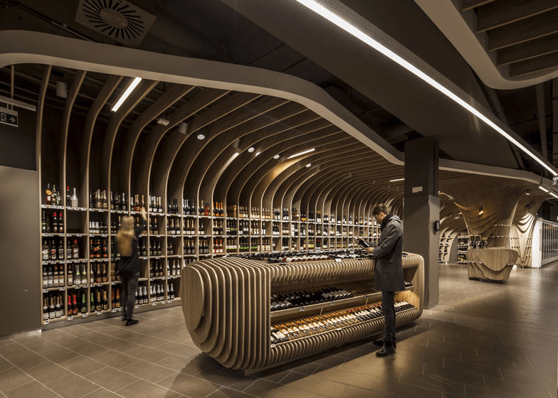 Interior de una tienda de vinos con estanterías curvadas y modernas, donde un cliente examina la mercancía. Las luces LED resaltan la arquitectura de madera y el ambiente elegante del espacio.