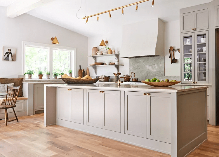 A modern kitchen featuring light gray cabinetry, a spacious island, wooden countertops, and various decorative items on shelves. Natural light streams in through large windows, complementing the earthy tones.