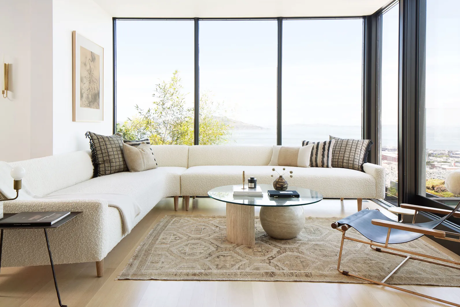 A modern living room featuring a large white sectional sofa with various decorative pillows, a round glass coffee table atop a patterned rug, and large windows providing a view of the outdoors.