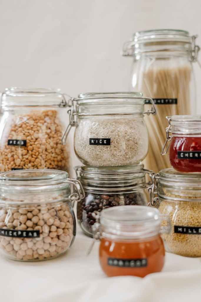 A collection of glass storage jars with labels, containing various grains and legumes, arranged neatly on a light surface.