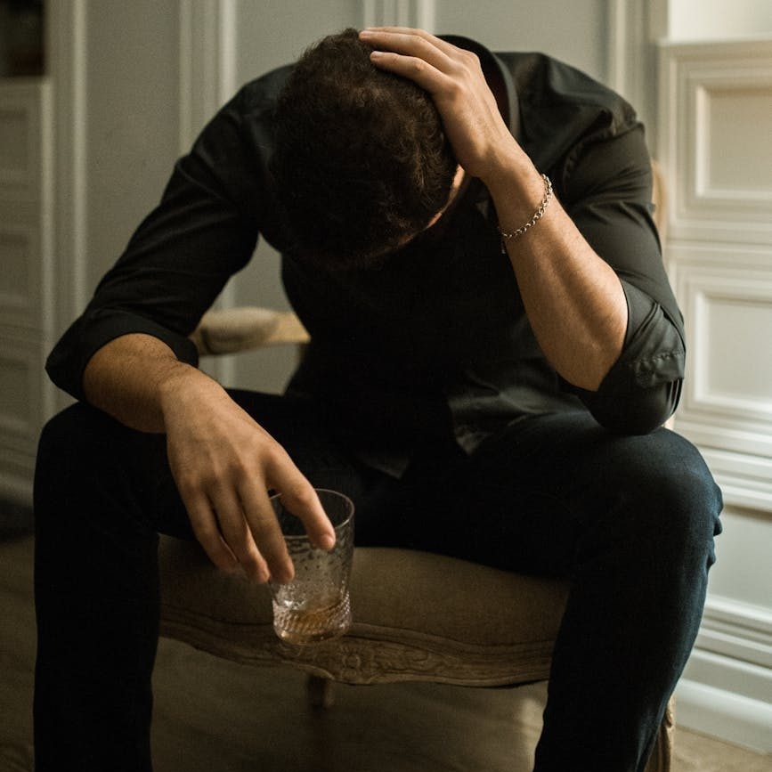 Un hombre sentado en una silla, cabizbajo y sosteniendo en una mano un vaso con licor y con la otra mano se toca la cabeza en seña de estrés y angustia. 
