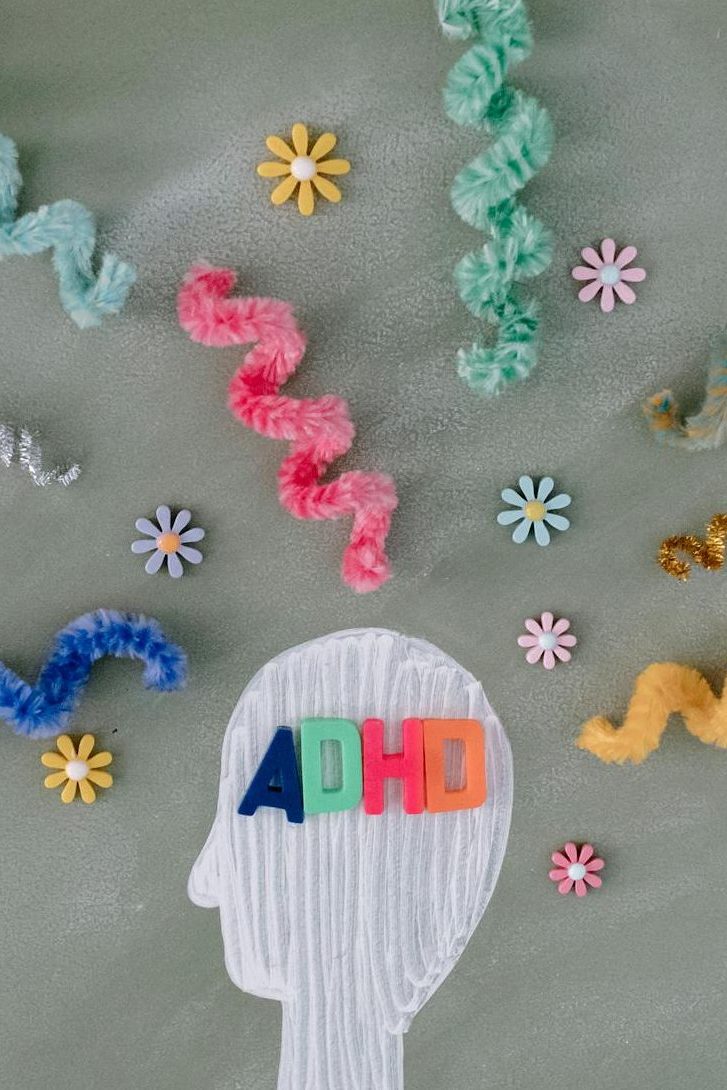 An artistic representation of a head silhouette with the letters 'ADHD' in colourful foam blocks, surrounded by wavy pipe cleaners and small flowers on a textured green background.