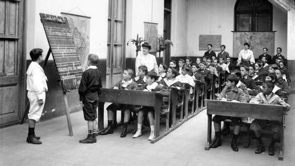 A historical black and white photograph of a classroom setting with a teacher, a chalkboard, and a group of young male students seated at wooden desks, attentively watching one student standing in front of the chalkboard.