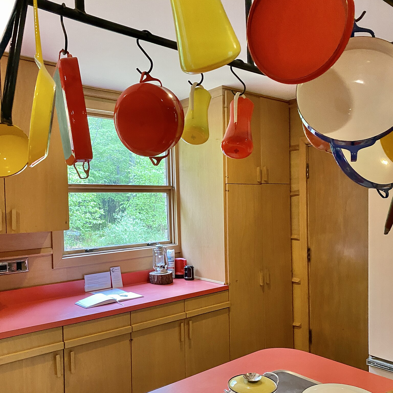 A bright kitchen with a red countertop and wooden cabinets, featuring an assortment of coloured pots and pans hanging from a rack above.