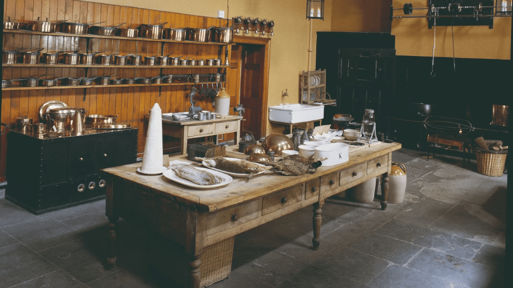 A historical kitchen featuring a large wooden table with various cooking utensils and ingredients, a black stove, and shelves lined with copper pots. The walls are painted a warm yellow, enhancing the rustic atmosphere.