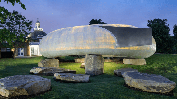A modern, oval-shaped sculpture perched on large stone pillars, illuminated under evening light. The sculpture is set in a grassy area with scattered rocks nearby, showcasing a blend of nature and contemporary design.