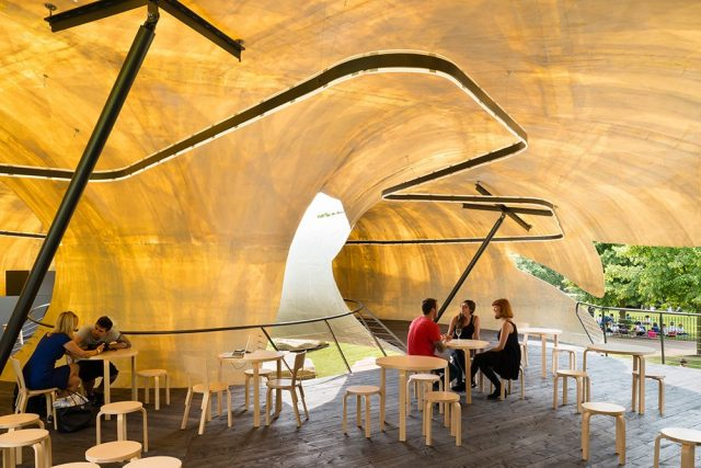 Interior of a modern, wavy café structure with large translucent walls, featuring a group of people sitting at tables and engaging in conversation.