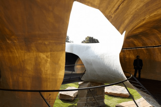 An interior view of a modern architectural space featuring curved walls and organic shapes, with a person standing in the background. Natural light filters through openings, illuminating stone elements and greenery.