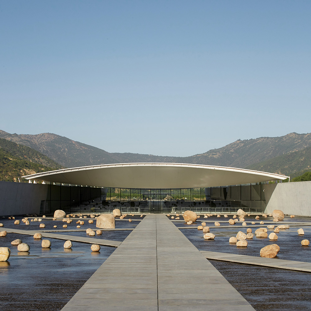 A modern architectural building with a curved roof, located by a body of water, surrounded by large stones and mountains in the background.