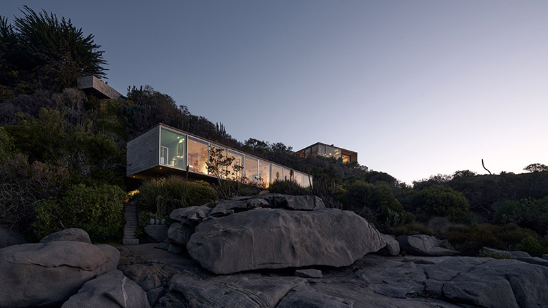 A modern glass house set against a rocky landscape during twilight, surrounded by greenery and a serene atmosphere.