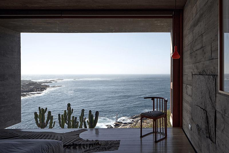 Interior view of a modern room with a large window overlooking the ocean, featuring a wooden floor, a cactus plant, and a stylish chair.