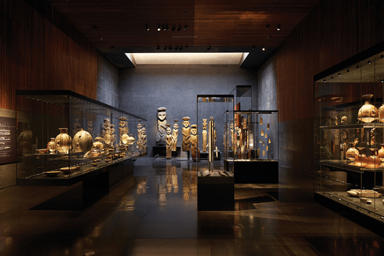 Interior of a modern museum showcasing various artifacts in glass display cases, including ancient sculptures and pottery, with wooden paneling and ambient lighting.
