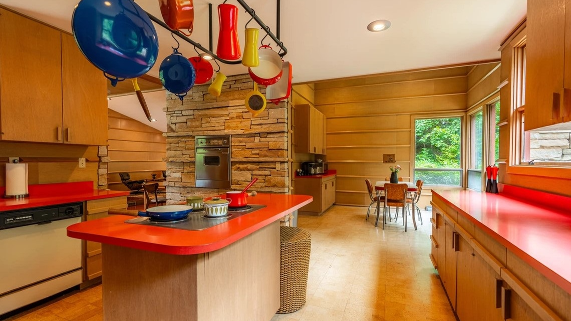 Bright and modern kitchen featuring a red countertop, wooden cabinetry, and stone accent wall. Hanging pots and pans add a splash of colour, with a dining area visible through large windows.